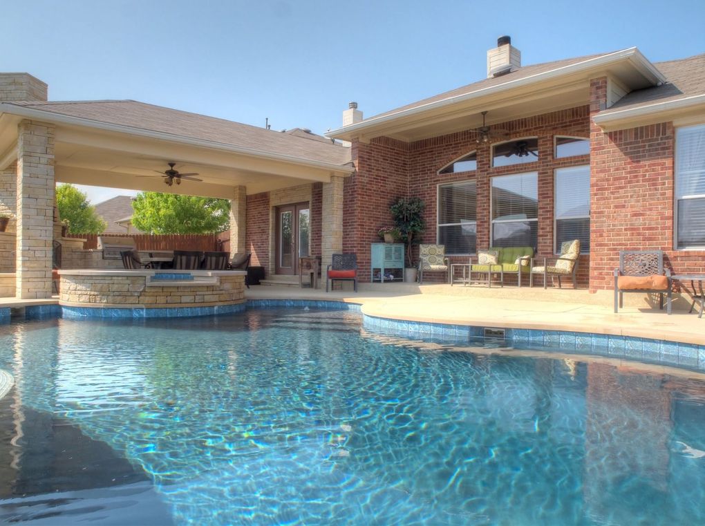 Backyard swimming pool beside a covered patio and brick house with blue water and stone coping