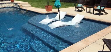 Poolside lounge chairs partly submerged in a blue swimming pool beside tan tiles and patio furniture