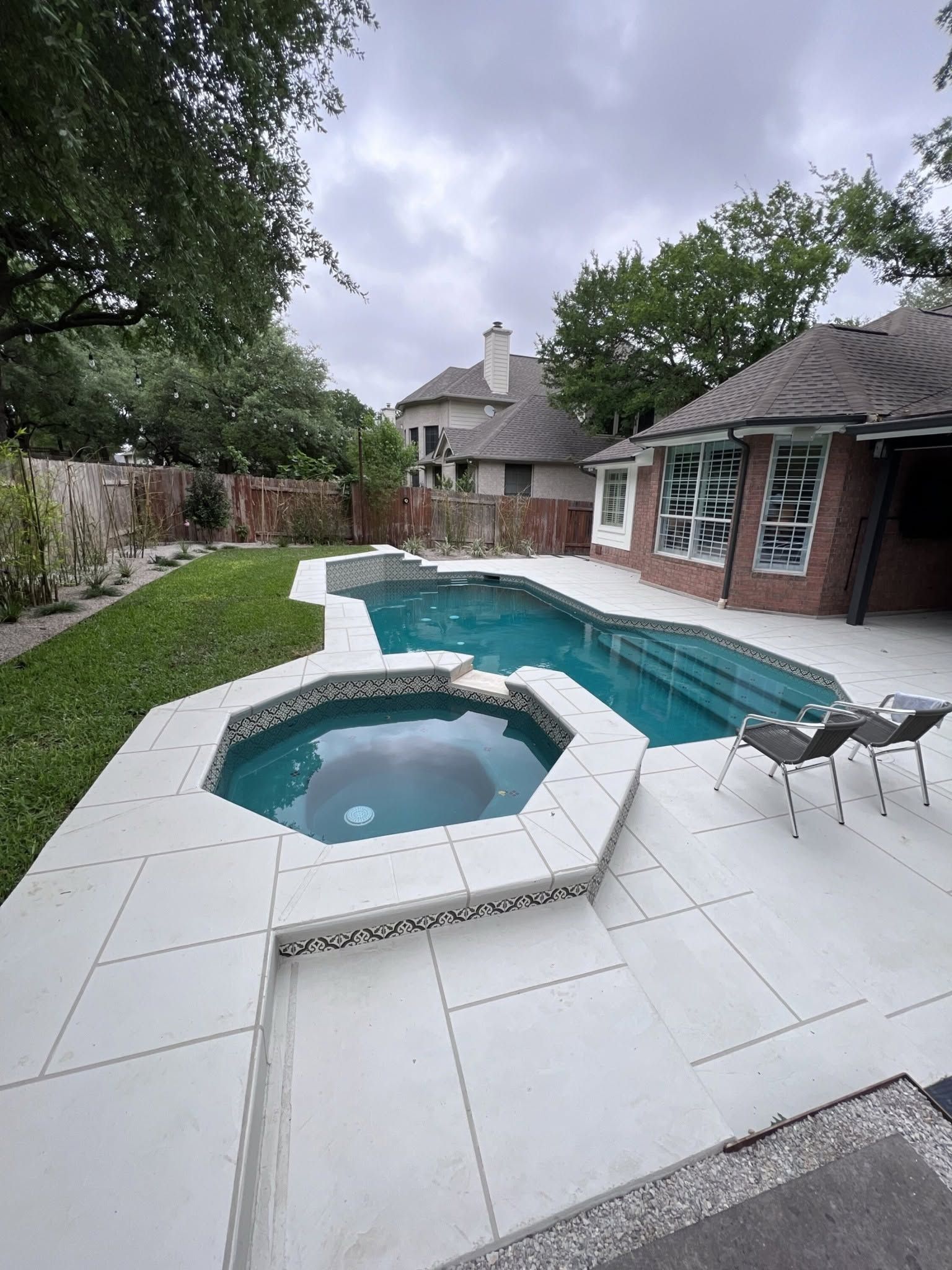 Backyard with white patio, turquoise pool and spa, brick house, and lounge chairs under cloudy sky