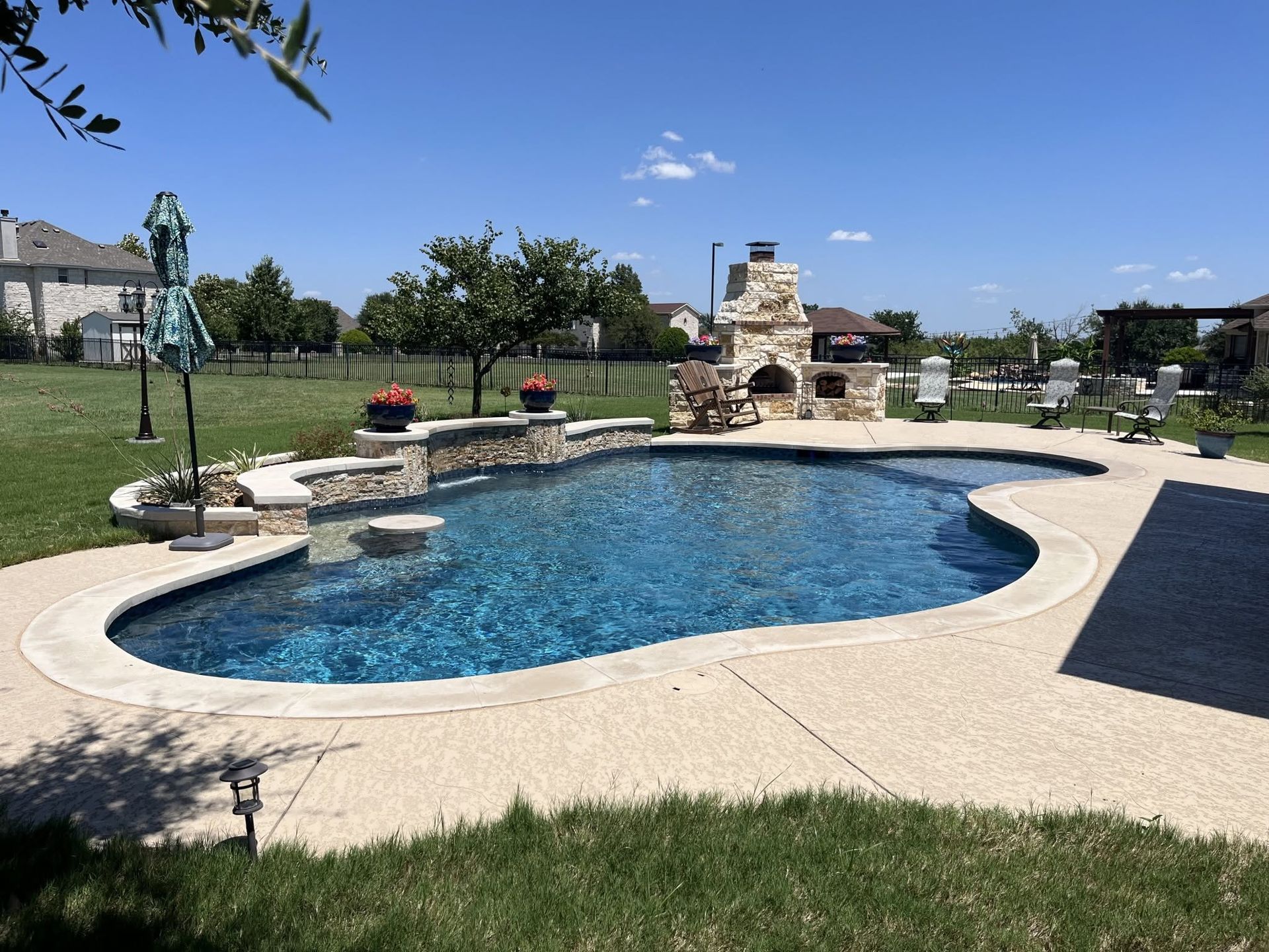 Backyard pool with curved stone deck, waterfall fountain, and lounge chairs under a clear blue sky