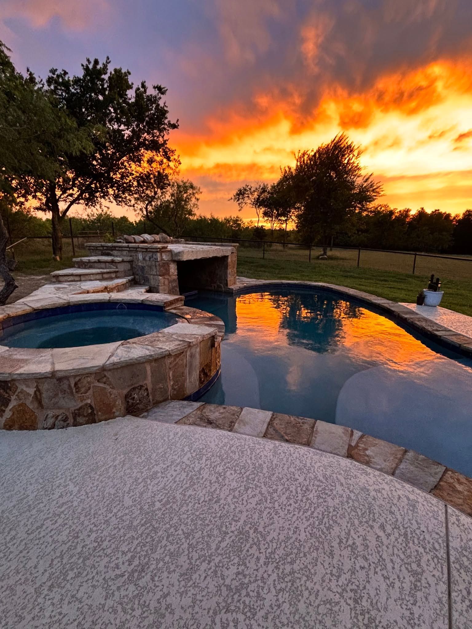 Backyard pool and stone hot tub at sunset, with orange clouds reflecting on the water.