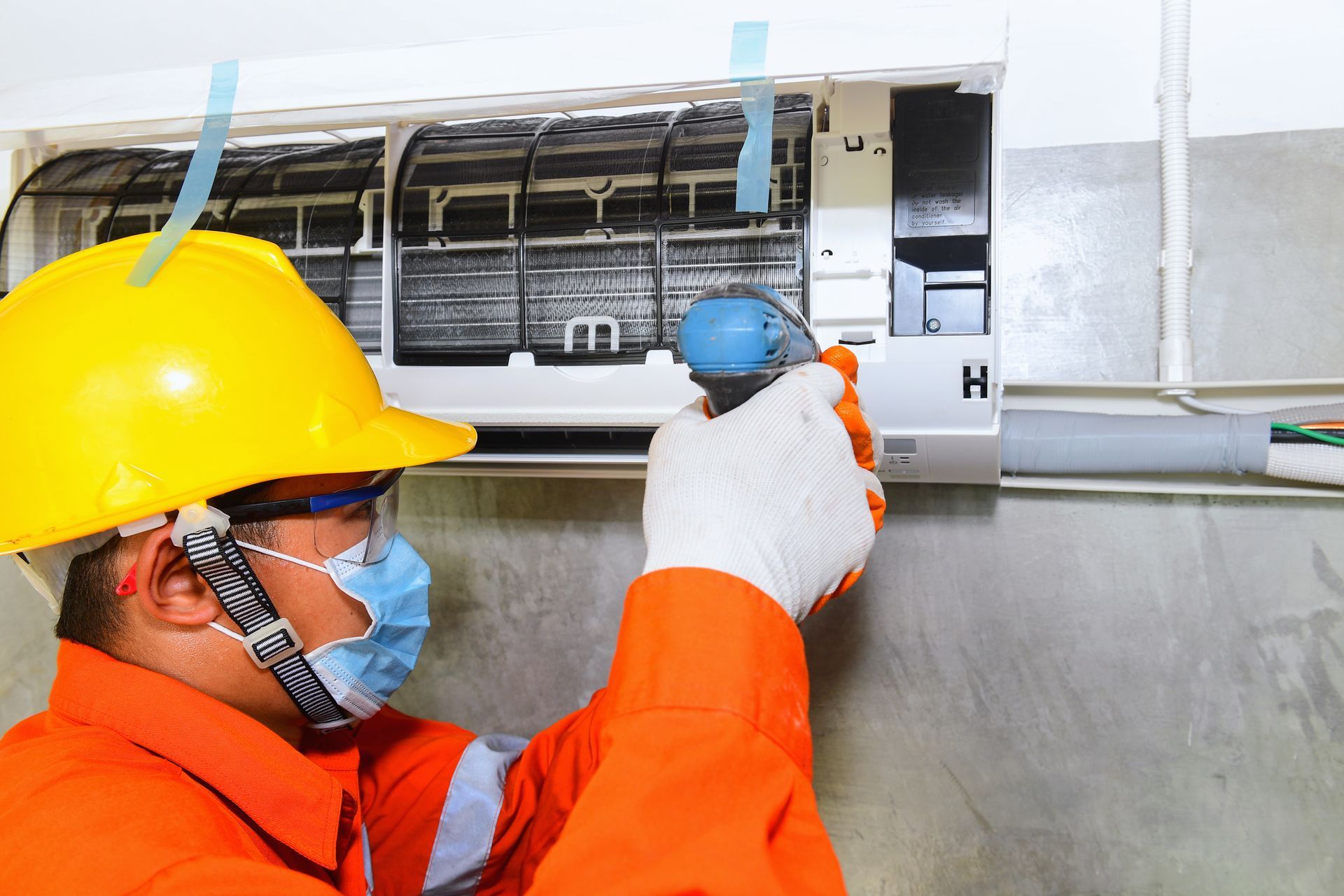Technician in protective gear performing air conditioning repair on an indoor wall-mounted AC unit.
