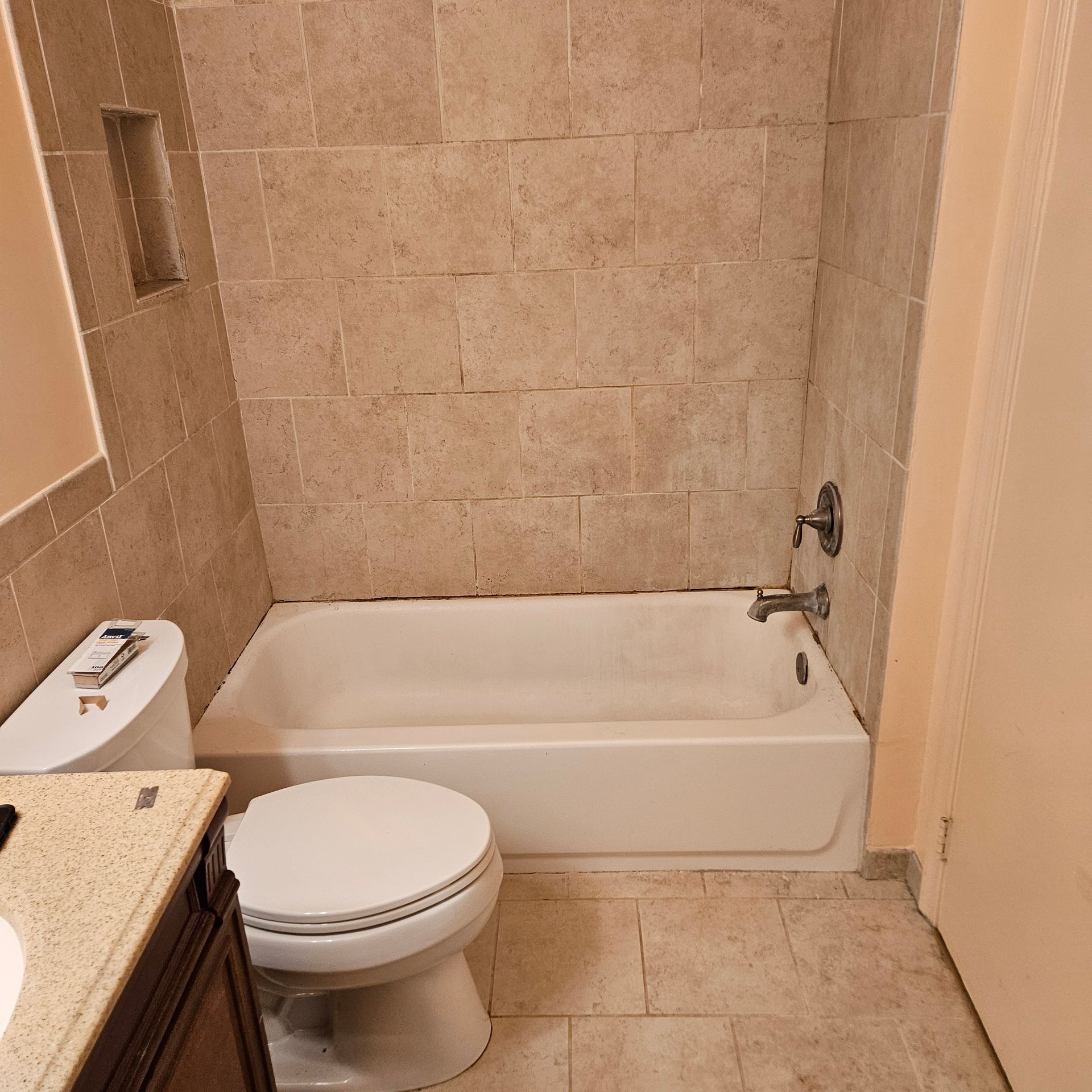 Small beige-tiled bathroom with a toilet, bathtub, and granite sink counter.