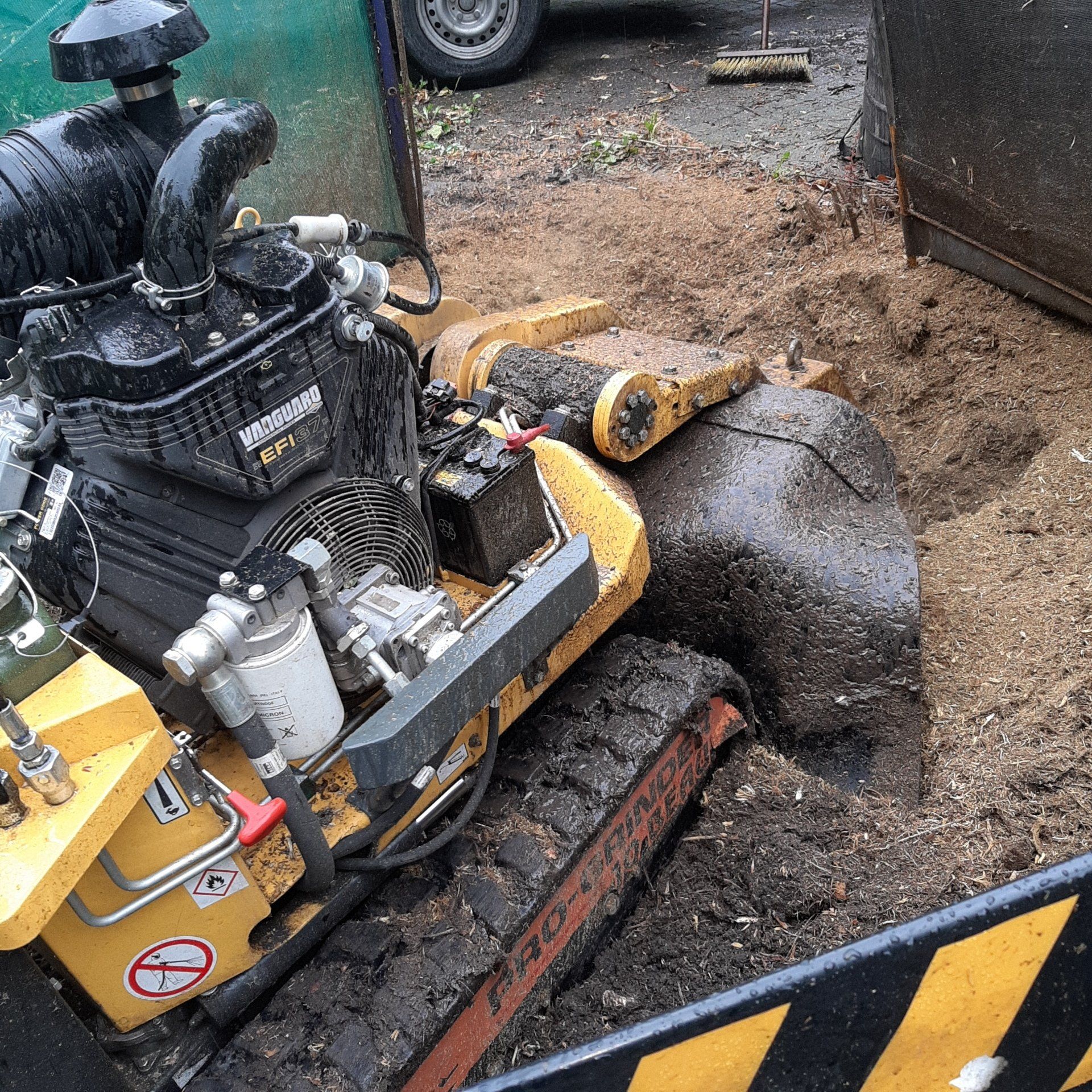 Geel-zwarte rupsbandmachine in een modderige opgraving. Motor zichtbaar, vlakbij puin.