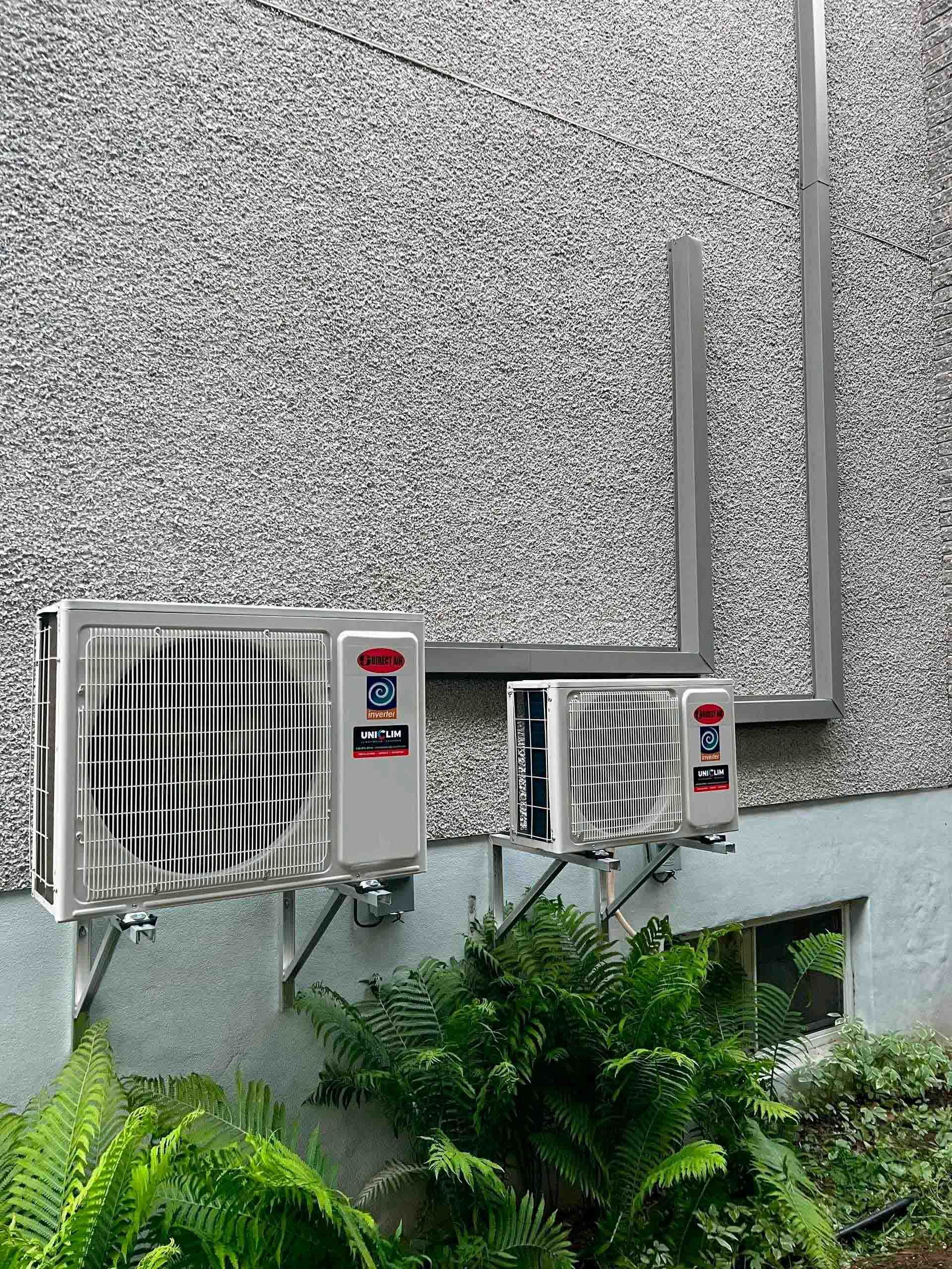 Deux unités de climatisation fixées sur un mur extérieur gris gravillonné, au-dessus d'une plante verte feuillue.