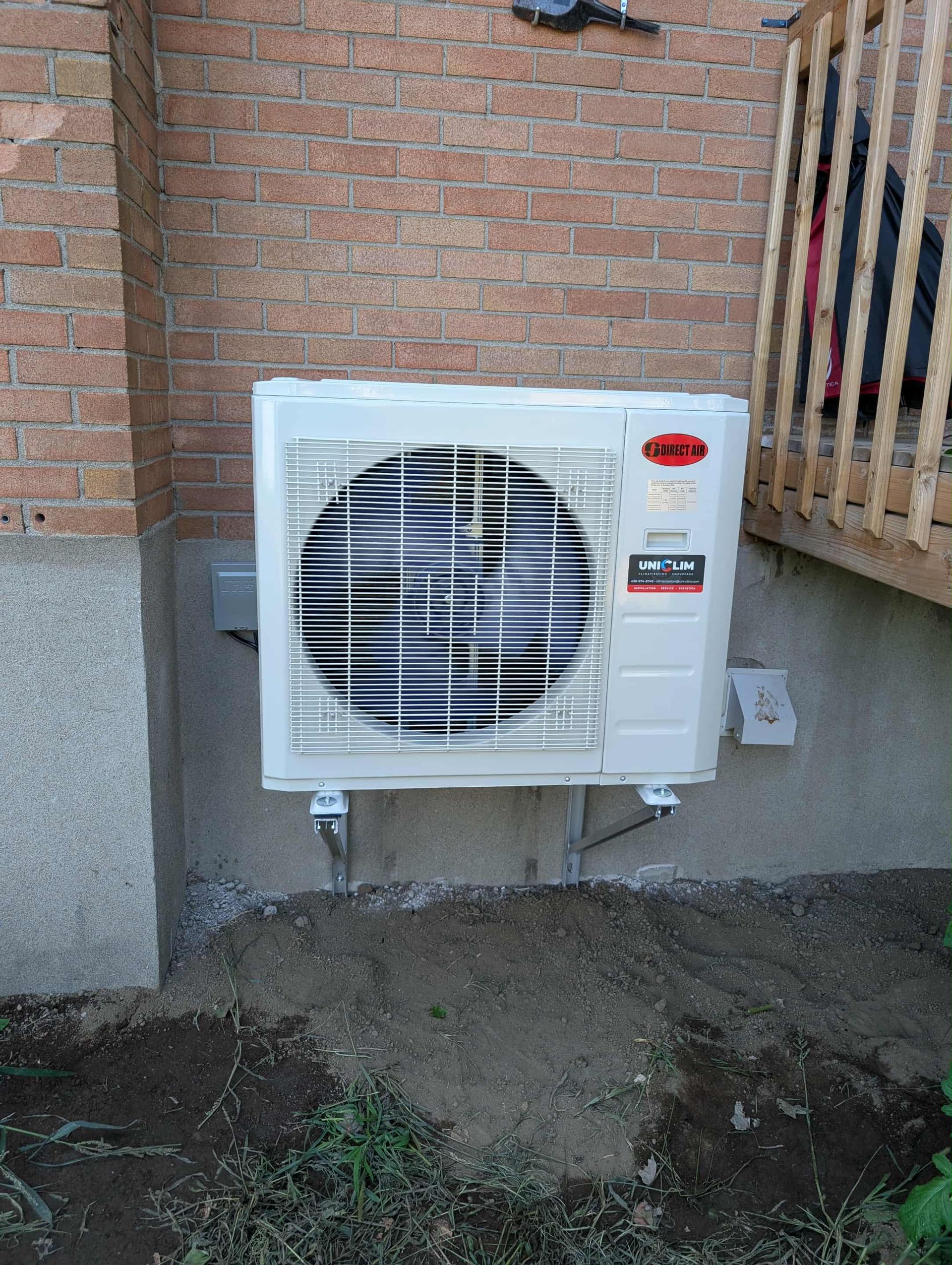 Un climatiseur ou une pompe à chaleur extérieure blanche, fixée sur un mur de sous-sol en pierre, à côté d'une terrasse en bois.