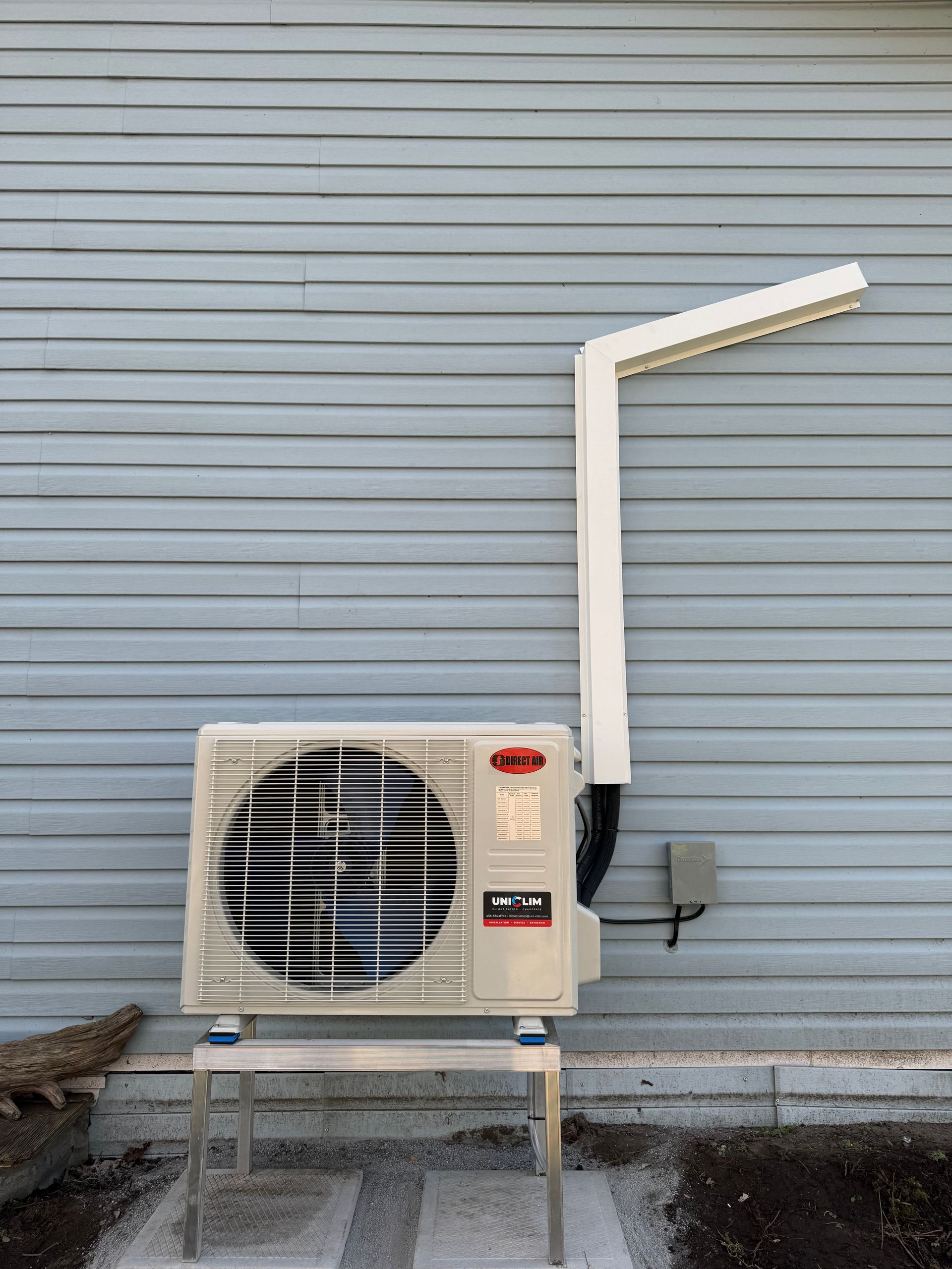 Une unité de pompe à chaleur extérieure mini-split surélevée sur un support métallique contre un revêtement gris clair avec un couvercle de conduit mural blanc.