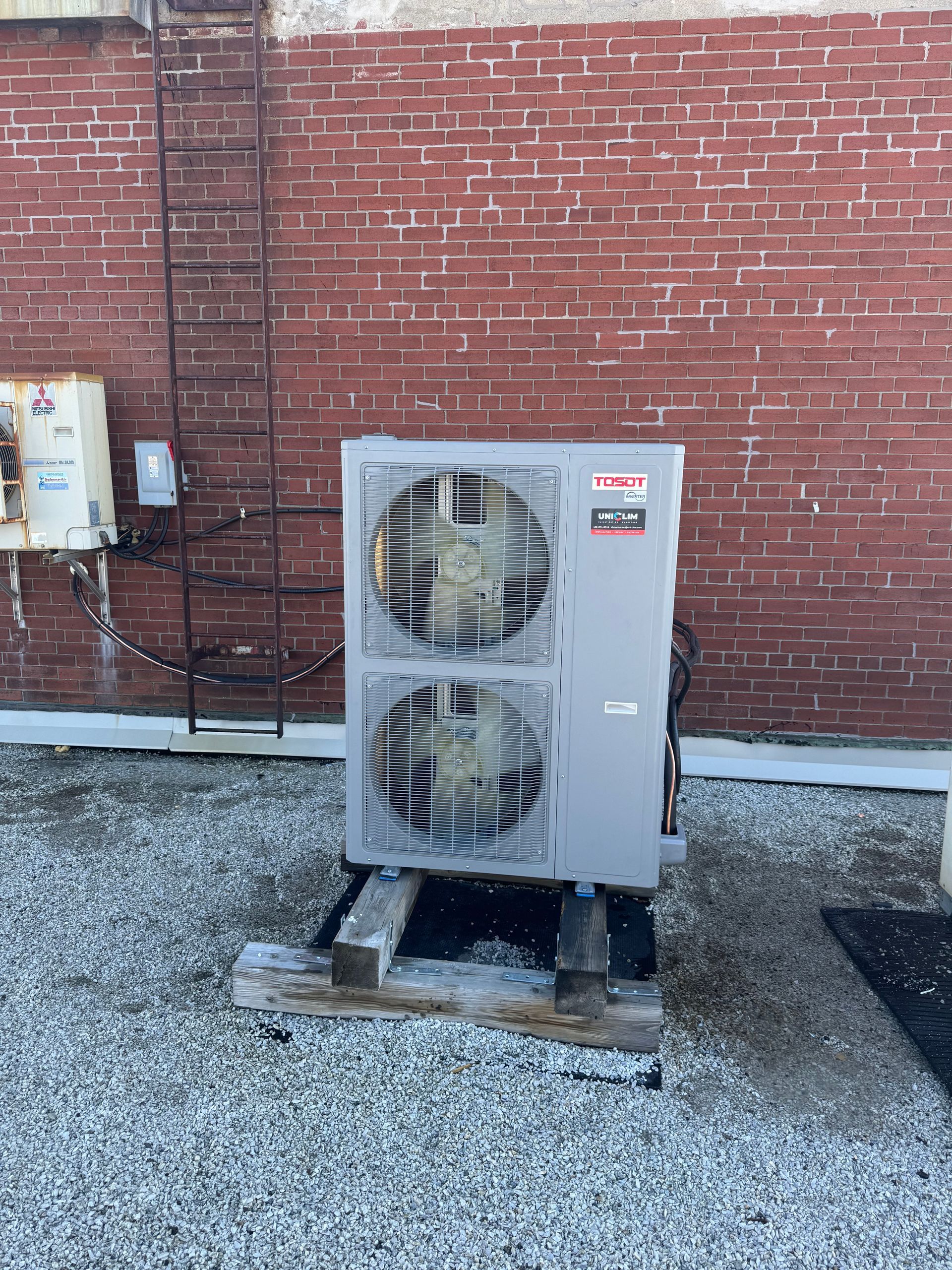 Un appareil de climatisation gris à deux ventilateurs est posé sur des palettes en bois, sur une surface gravillonnée, devant un mur de briques rouges avec une échelle métallique.