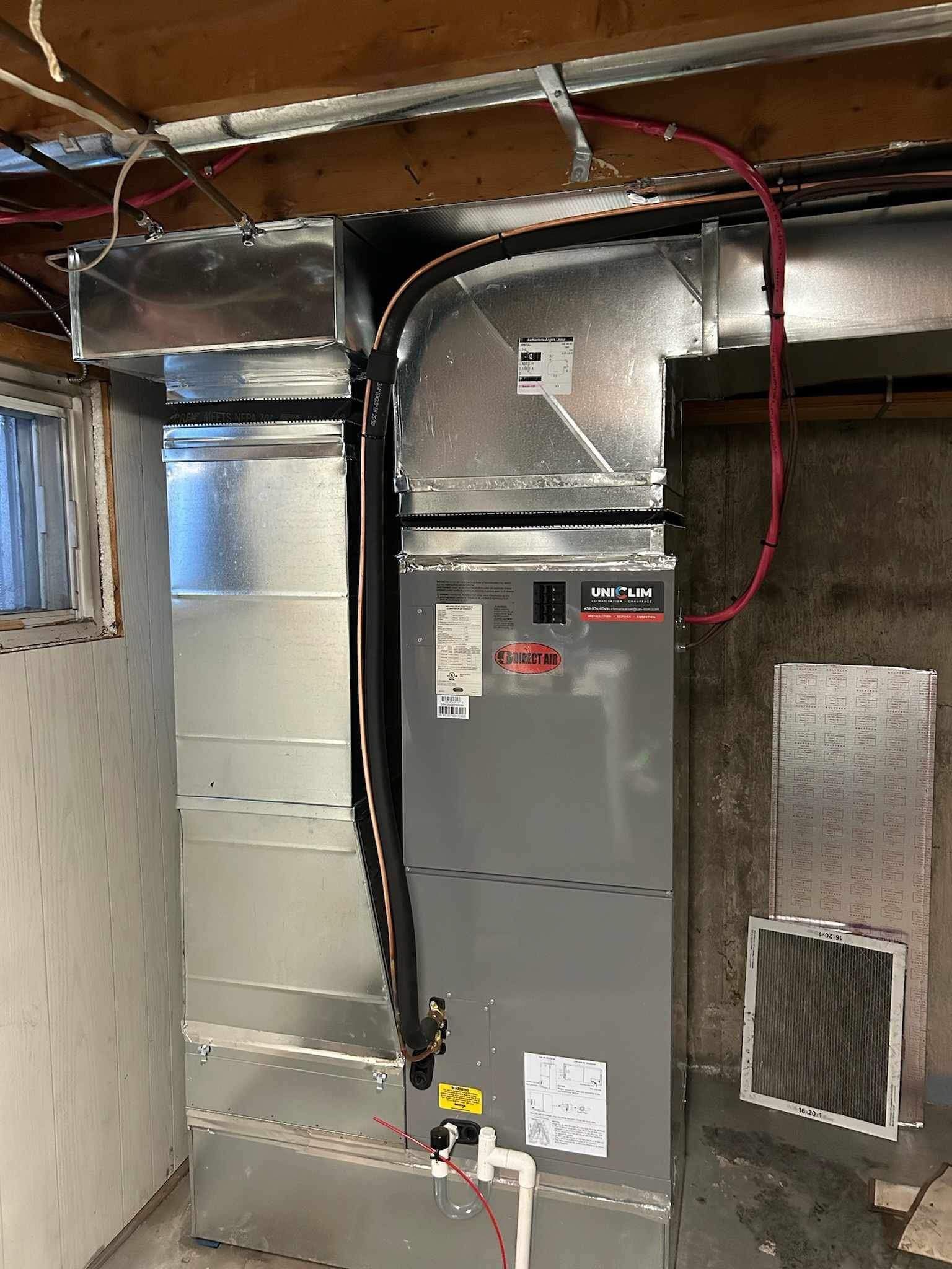 Une unité de chauffage, ventilation et climatisation grise installée contre un mur de sous-sol en béton, avec des conduits métalliques et des câbles apparents.