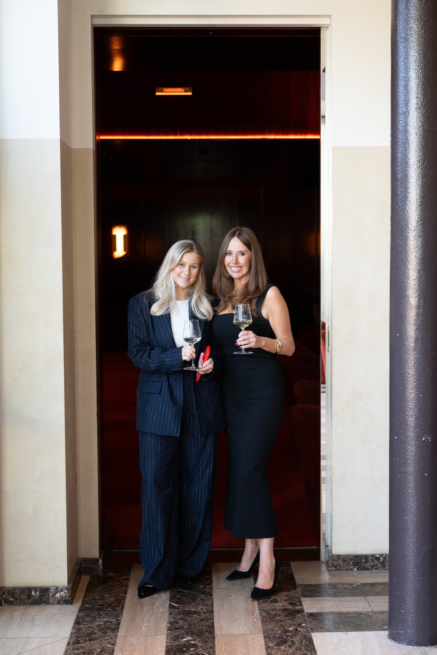 Two people in formal attire hold wine glasses while posing in a dark-lit doorway.