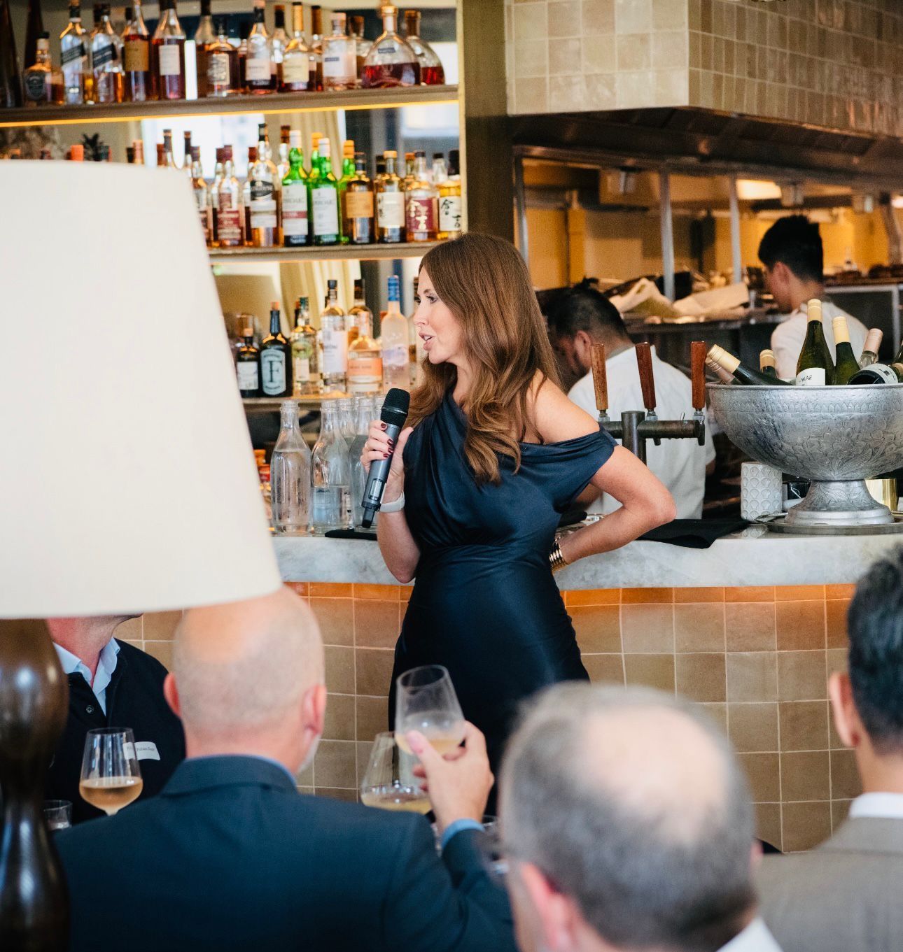 A person in a dark, off-the-shoulder dress speaks into a microphone at a bar while guests listen in the foreground.
