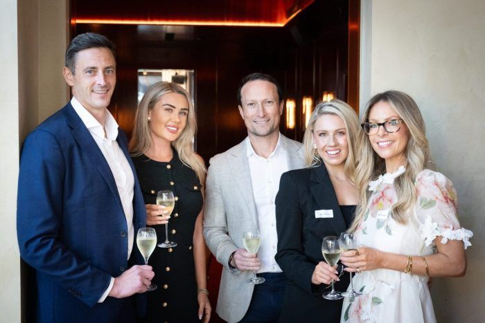 Five colleagues holding champagne flutes stand together in a well-lit, professional setting.