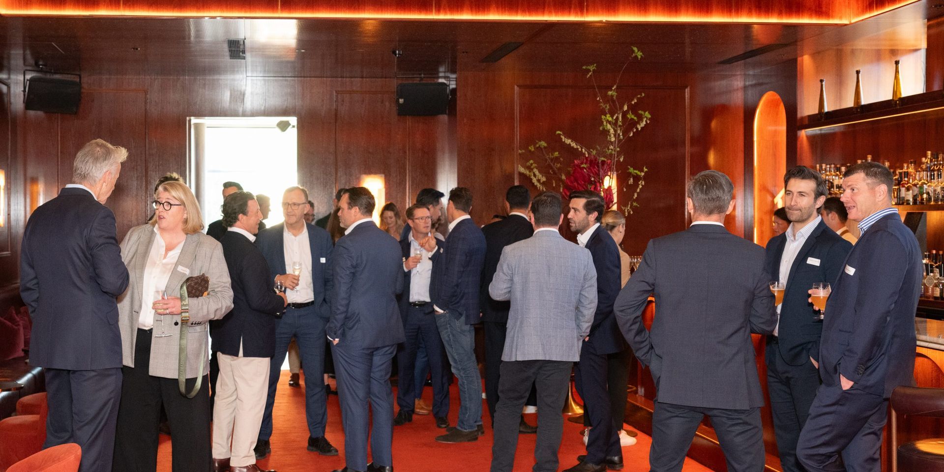 A group of professionals in suits and formal wear socialize in a dimly lit, wood-paneled room with warm lighting.