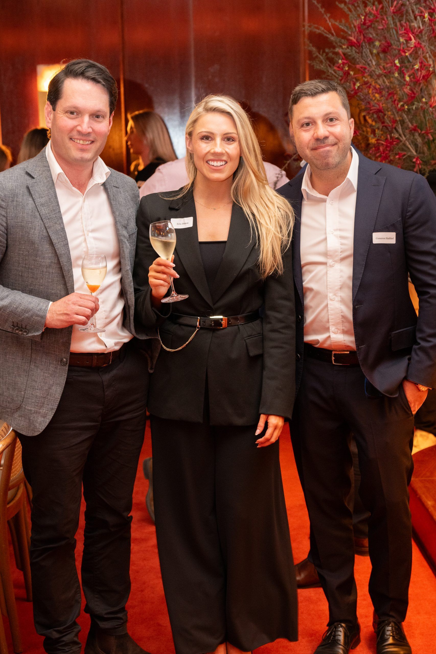 Three people in professional attire pose for a photo at an indoor event, holding glasses and smiling.