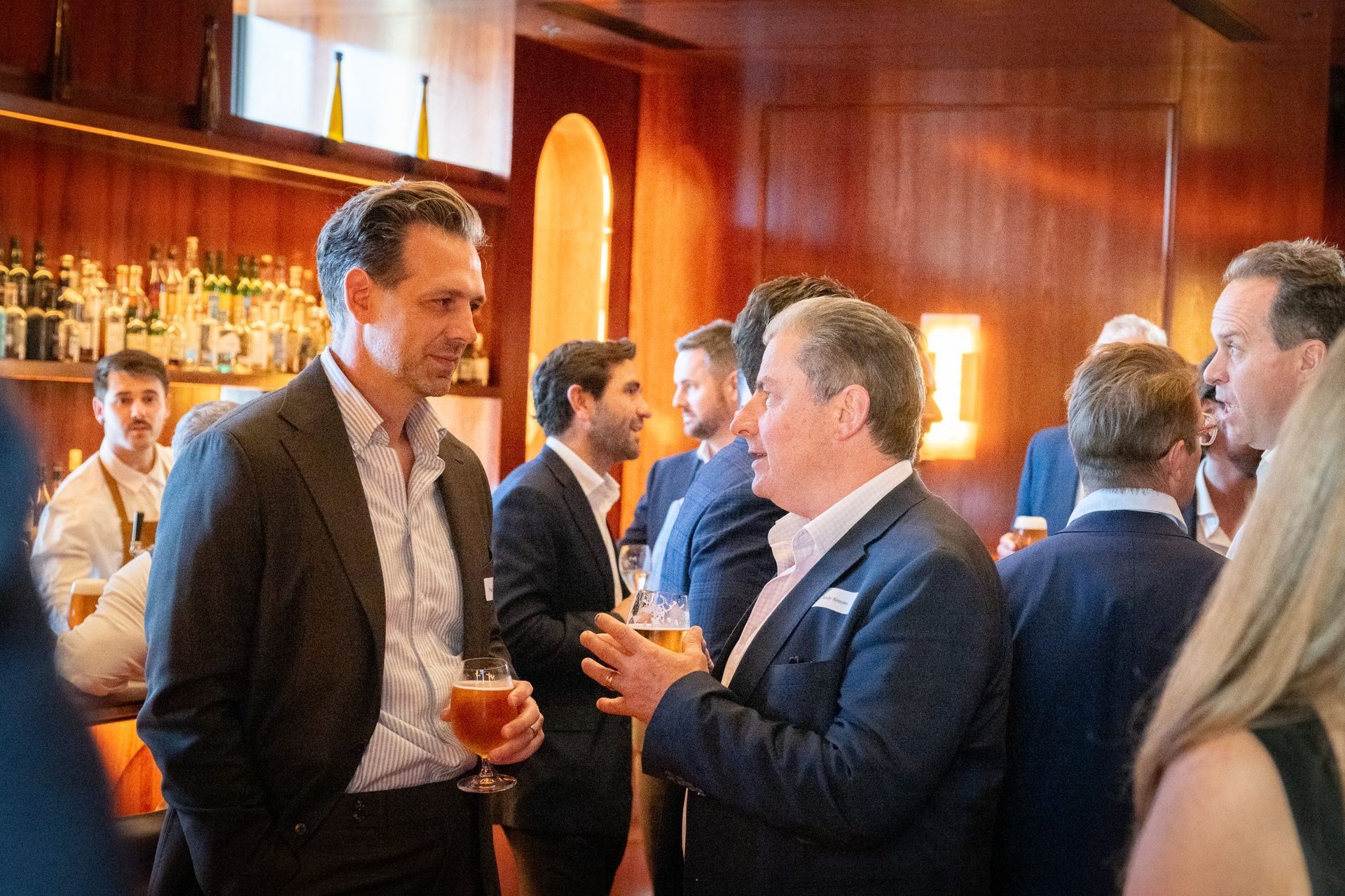 People in business attire chat and hold drinks at a social gathering in a room with wood-paneled walls.