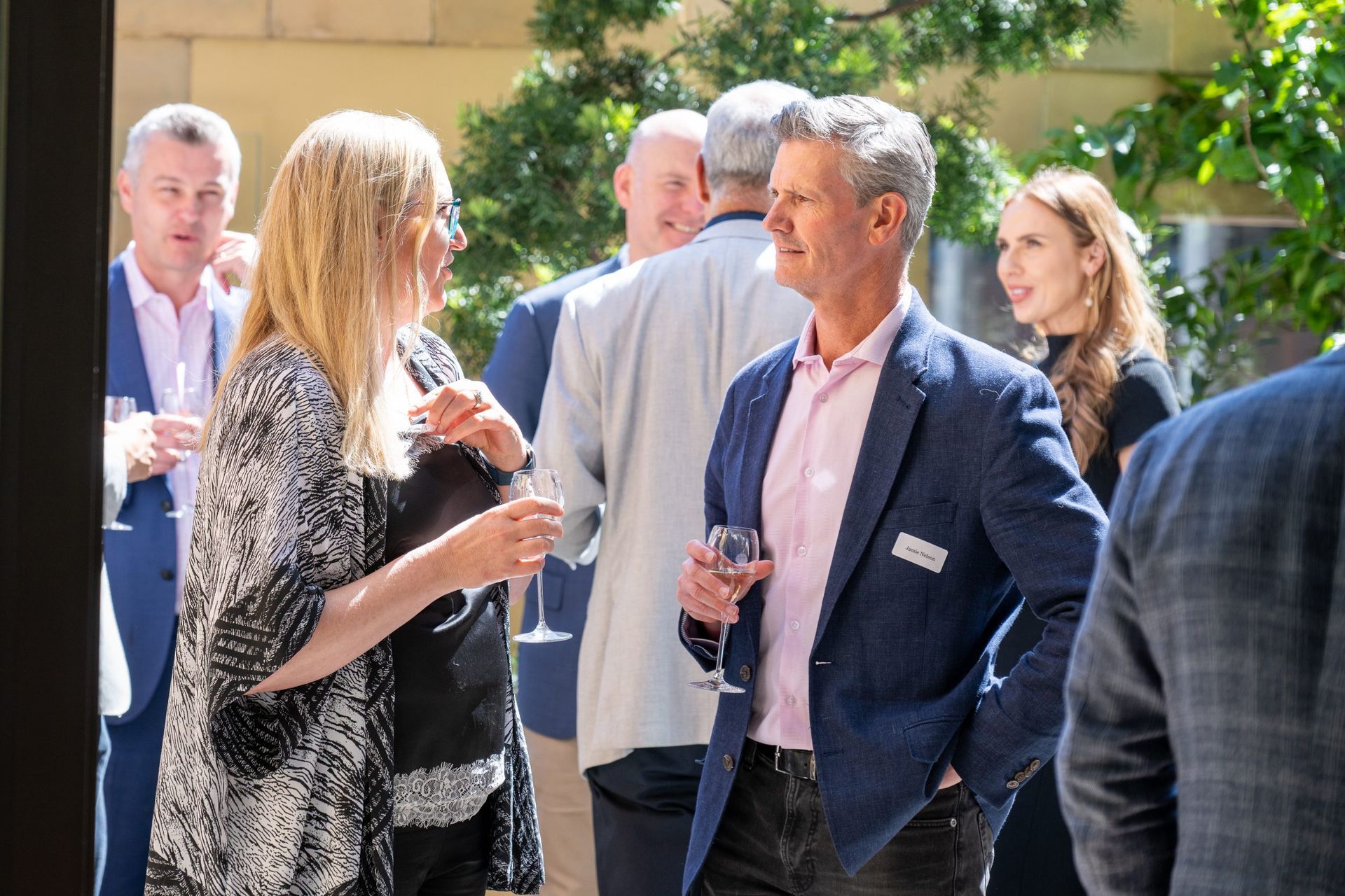 Attendees socialize outdoors at an event, holding drinks and chatting in a sunlit garden setting.