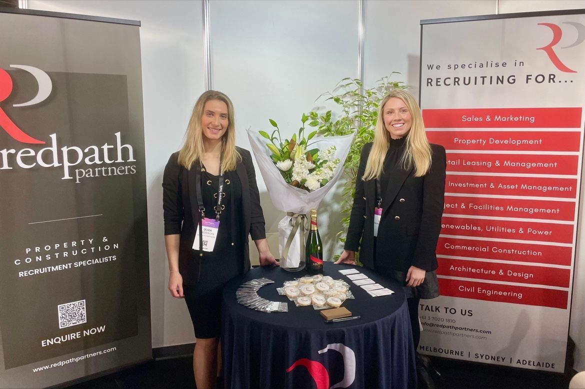 Two representatives stand at a branded Redpath Partners display booth with a table of promotional materials and flowers.
