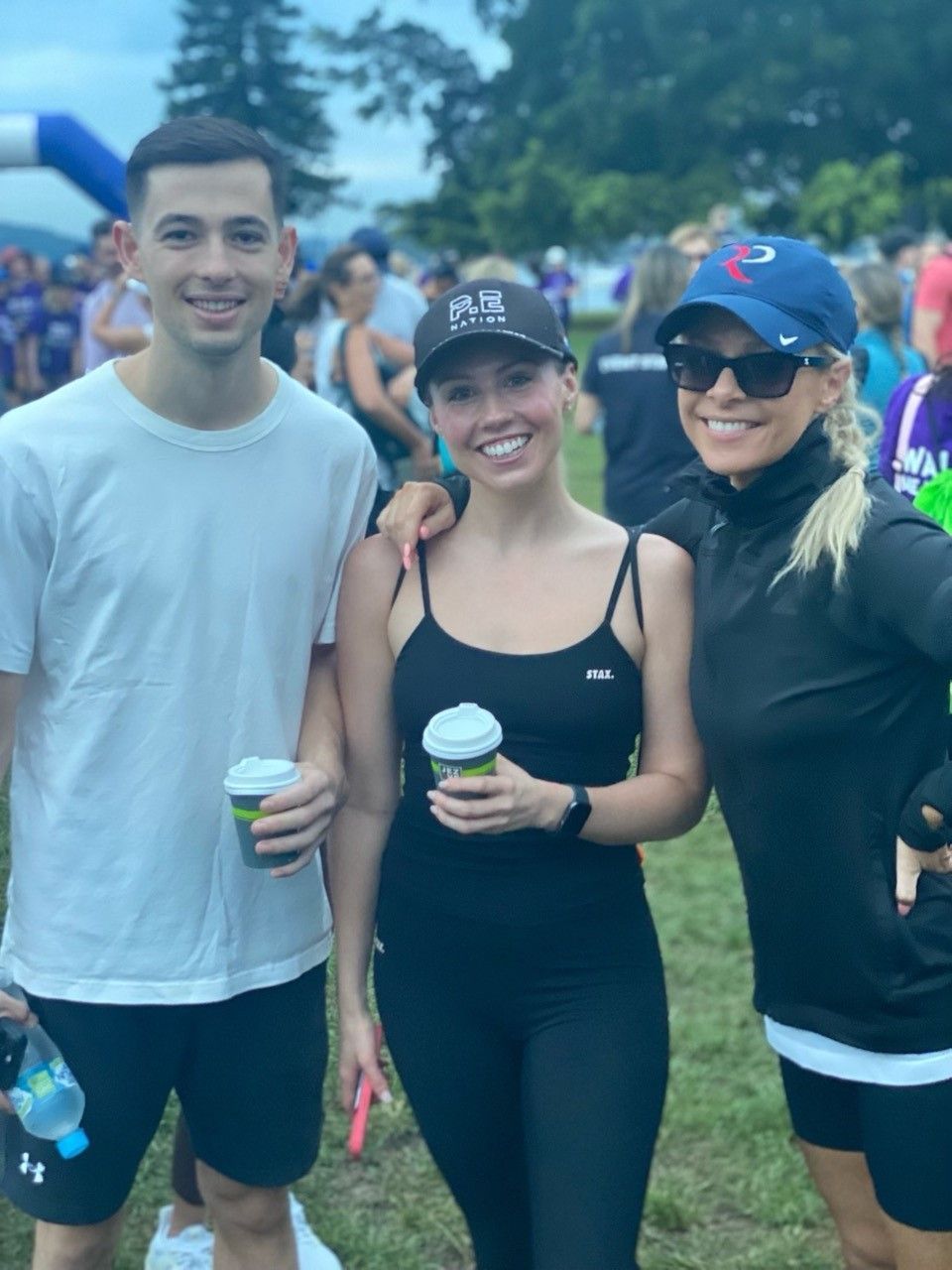 Three people standing together at an outdoor event, smiling and holding cups, with blurred crowds and trees in the back.