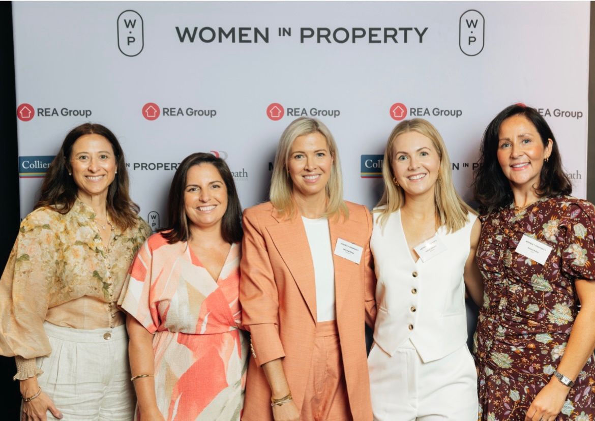 Five people stand smiling in front of a branded Women in Property banner at an REA Group event.