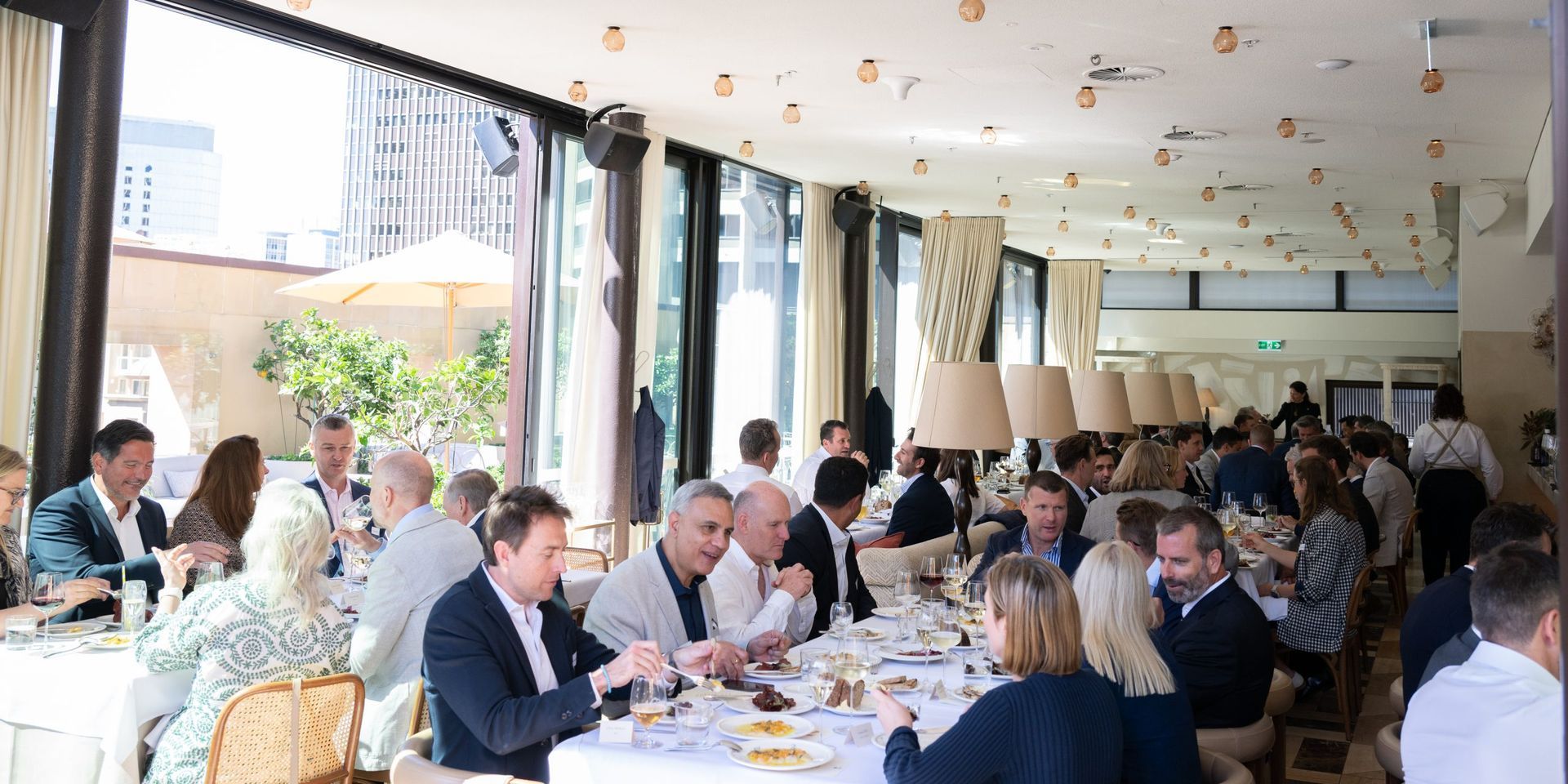 A brightly lit, modern indoor restaurant filled with people sitting at long, white-clothed tables, dining and talking.