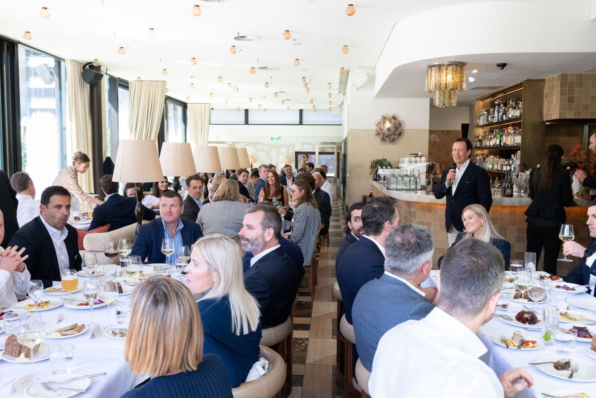 A group of people sitting at a long table in a bright, modern restaurant listening to a speaker standing near a bar.