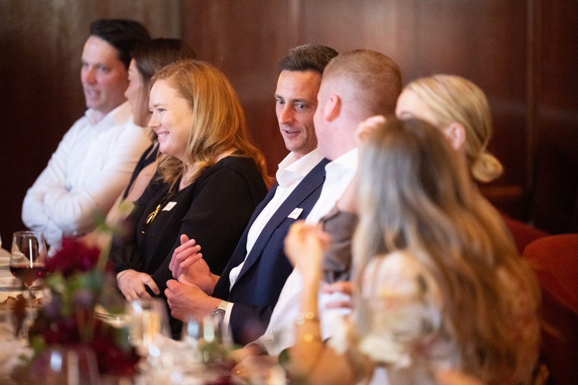 A group of people are seated at a dimly lit formal dinner table, talking and looking towards each other.