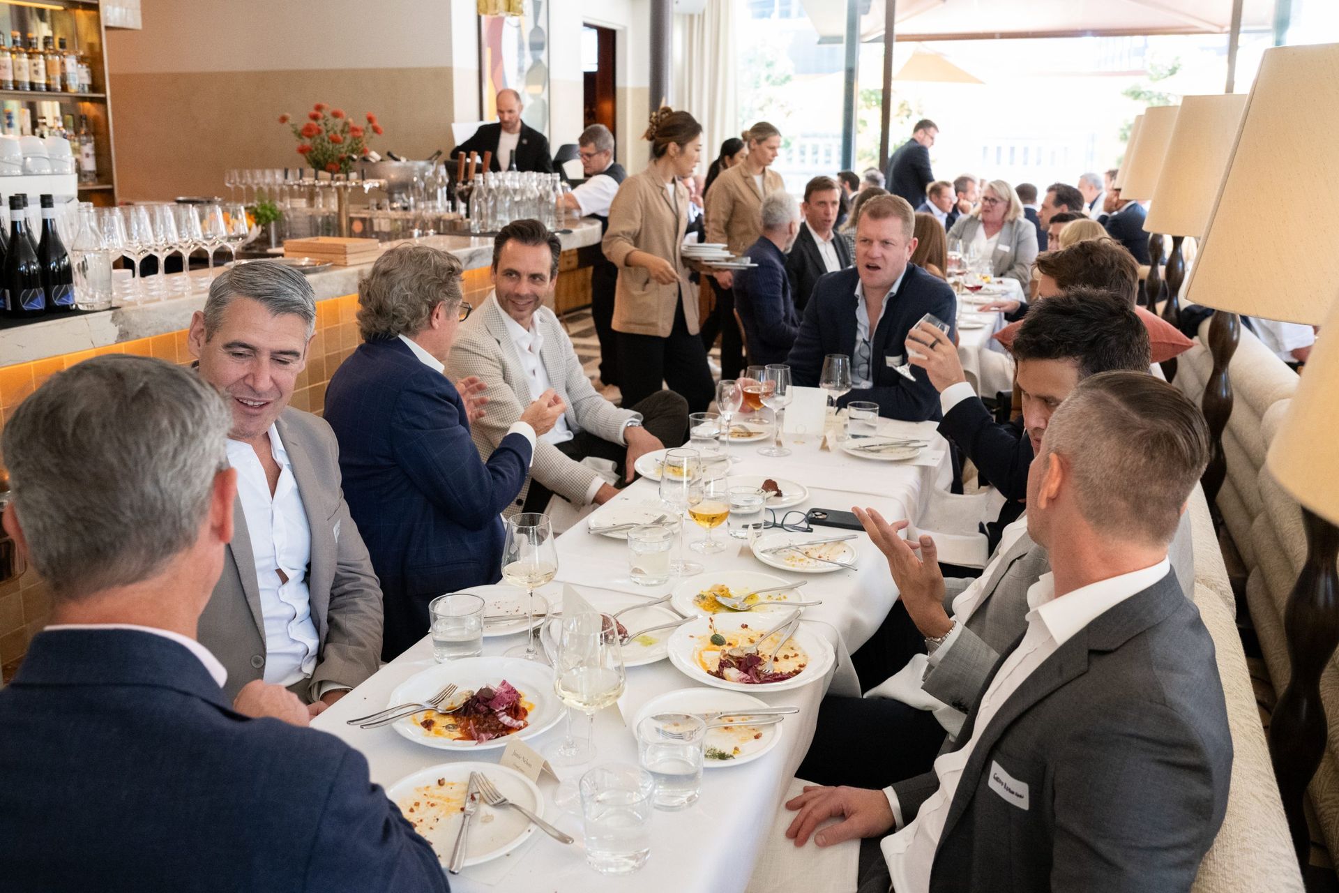 A group of professionals in suits dine and converse at a long table in a bright, upscale restaurant.