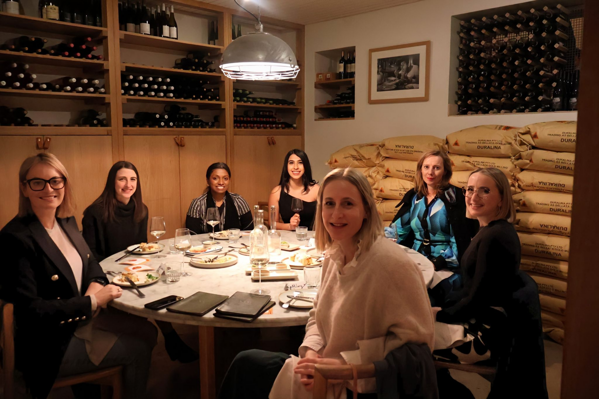Six people sit around a round dining table in a rustic room filled with wine bottles and stacks of grain sacks.