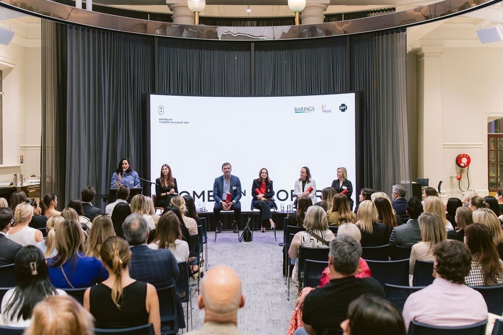 A panel of six people sits on a stage before an audience, with a white presentation screen in a formal event venue.