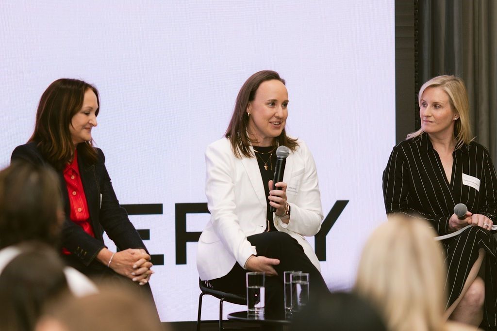 Three professionals sit on a stage during a panel discussion, with the center speaker holding a microphone.