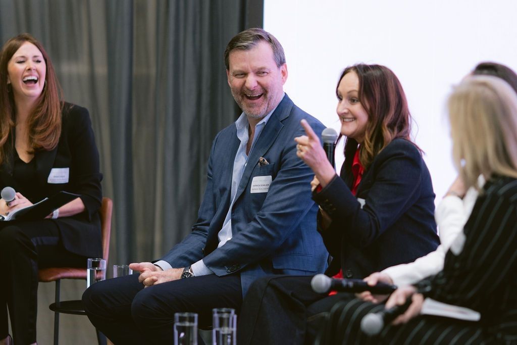 Four professionals seated in a row at a panel discussion, laughing and engaged in conversation with microphones.
