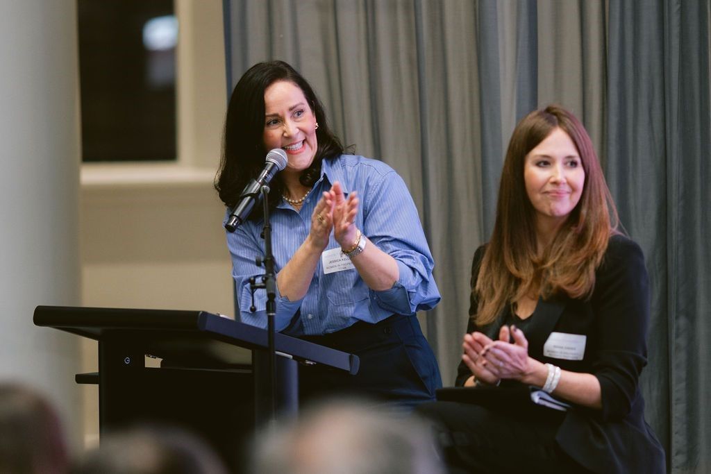 Two people sit at a podium and table, smiling and clapping, while facing an audience in a presentation setting.