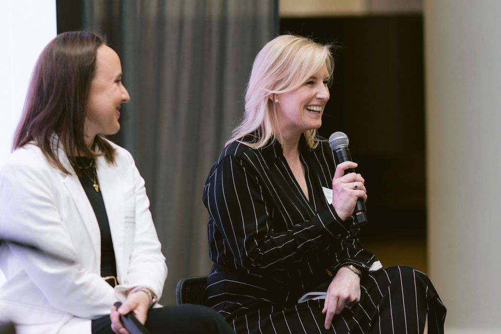 Two people sitting on a stage, one speaking into a microphone while the other listens and smiles.