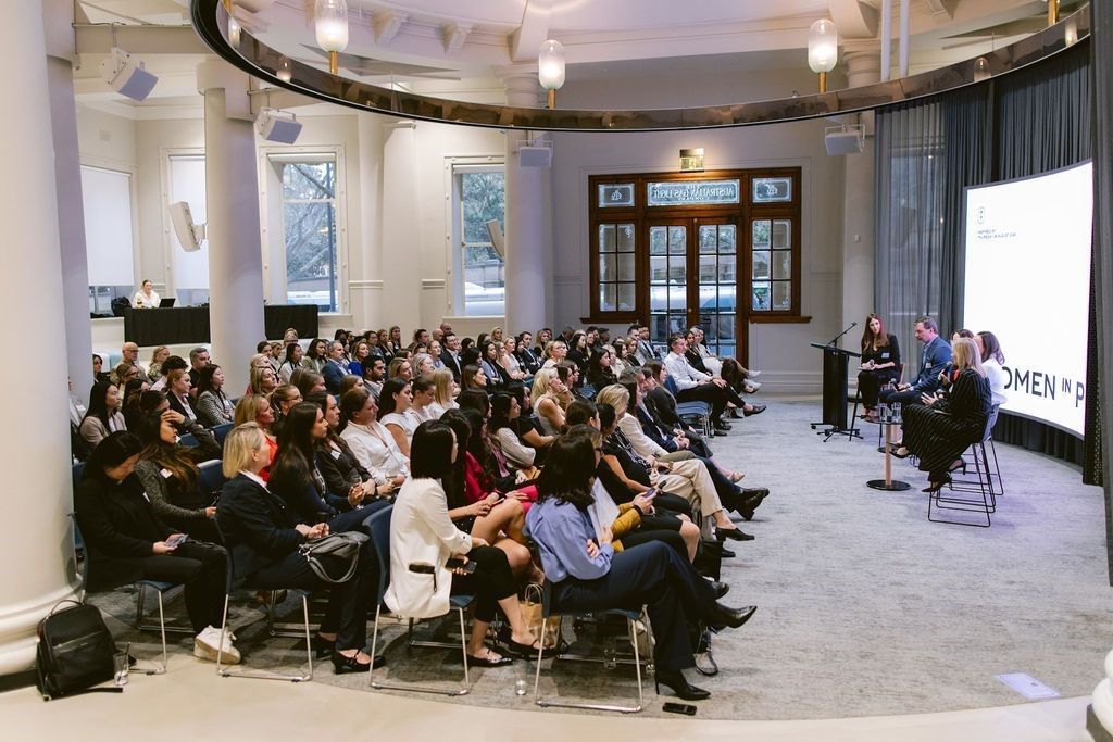 A large audience sits in an elegant, sunlit circular room, watching a panel discussion on a stage.