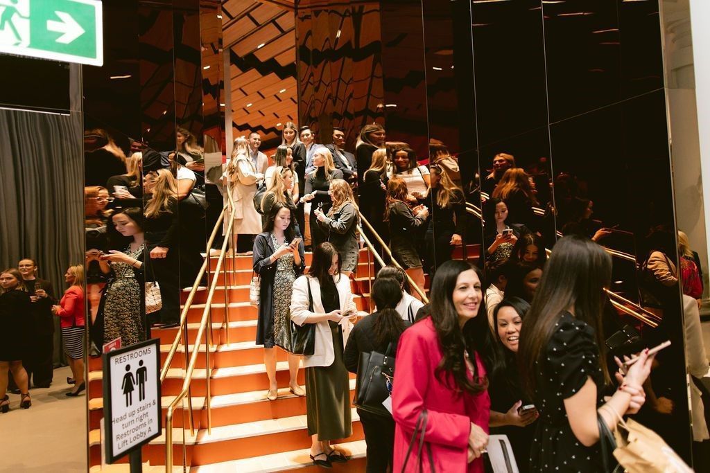 A crowd of people stands in a modern foyer on and around orange stairs, with many looking at their phones.