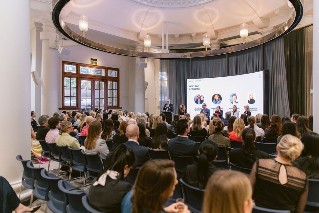 A panel discussion with speakers on a large screen facing an audience in a circular room with pillars and draped walls.