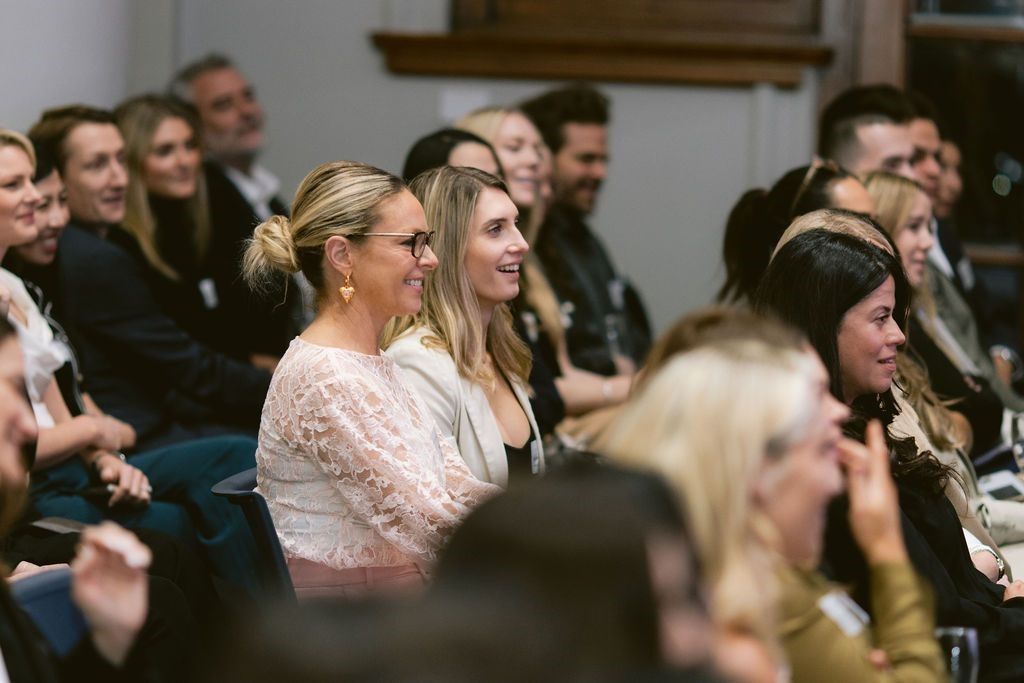 A group of people sitting in an indoor setting, listening attentively and smiling during a presentation.