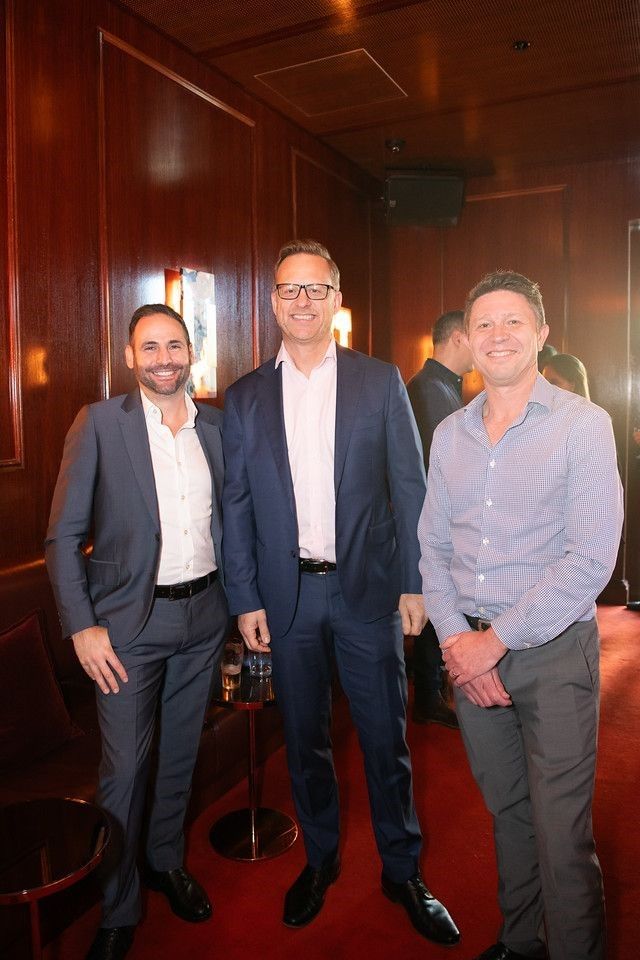 Three men in business-casual attire stand smiling together in a wood-paneled room with red carpeting.