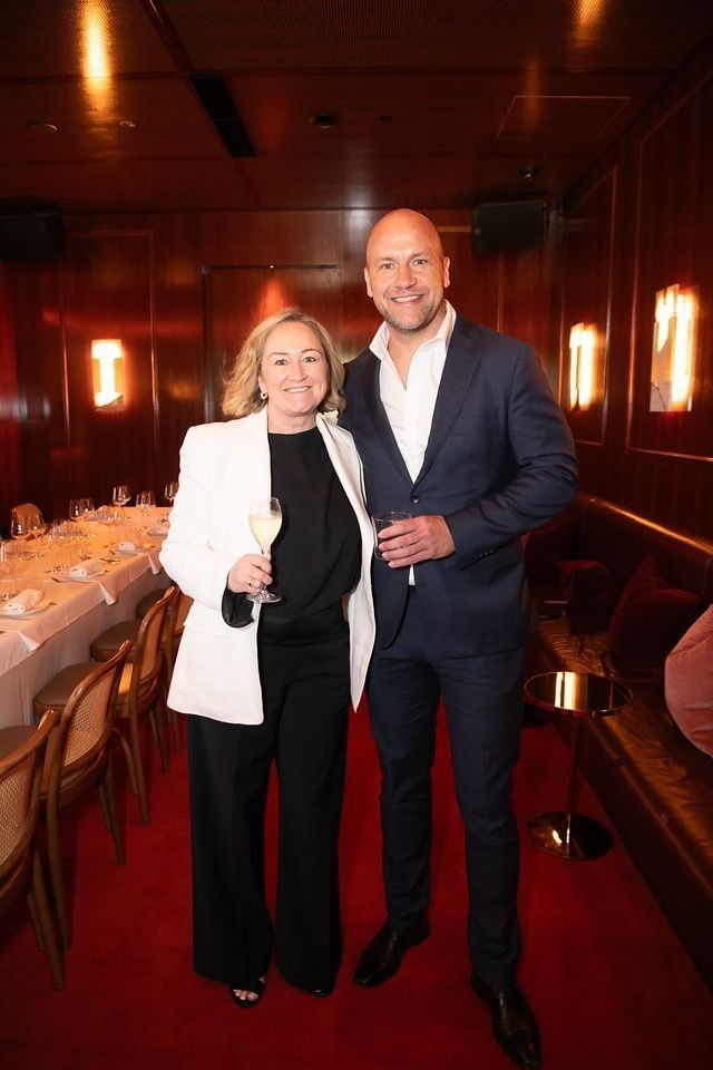 A smiling duo stands in a dimly lit, wood-paneled room with a long dining table, red carpet, and warm wall lighting.