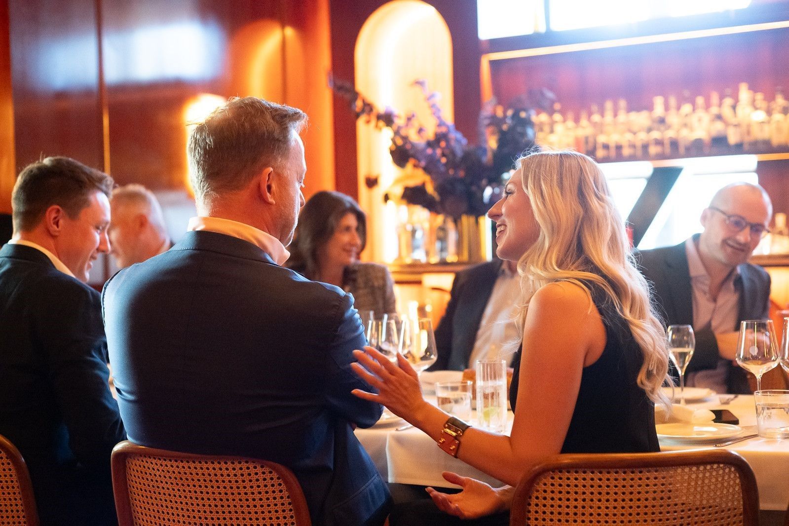 A group of people in business-casual attire dine at a table in a warm-lit restaurant while engaged in conversation.