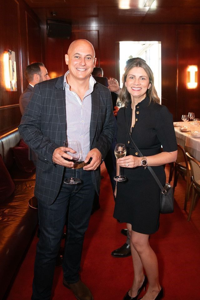 A man and woman in business casual attire smile while holding glasses of wine at an indoor evening event with red carpet.