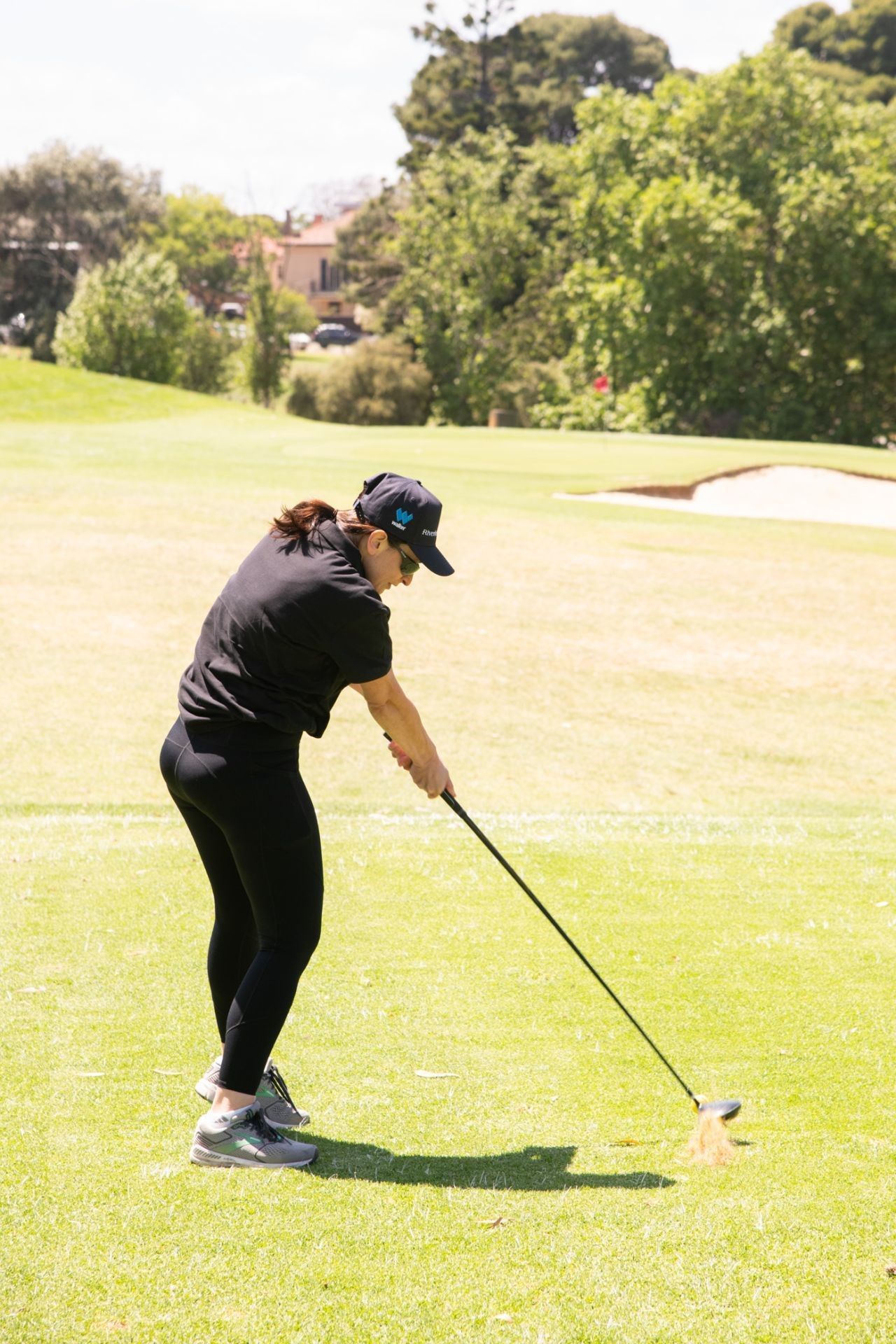 A person in black clothing hits a golf ball on a sunny green course with trees and a sand bunker in the background.