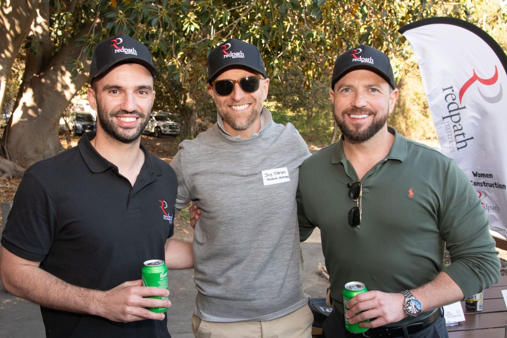 Three men in matching baseball caps smiling outdoors, two holding green cans, with a branded flag in the background.