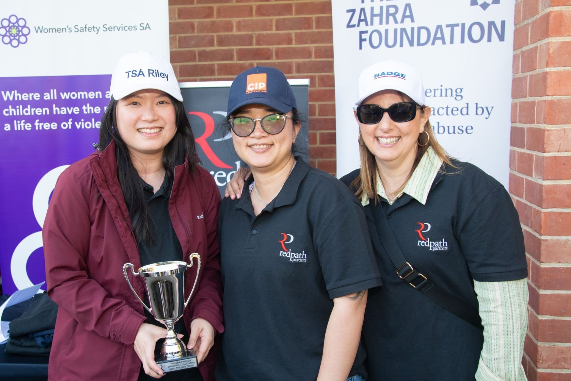 Three people wearing caps stand smiling in front of Zahra Foundation banners; the person on the left holds a trophy.