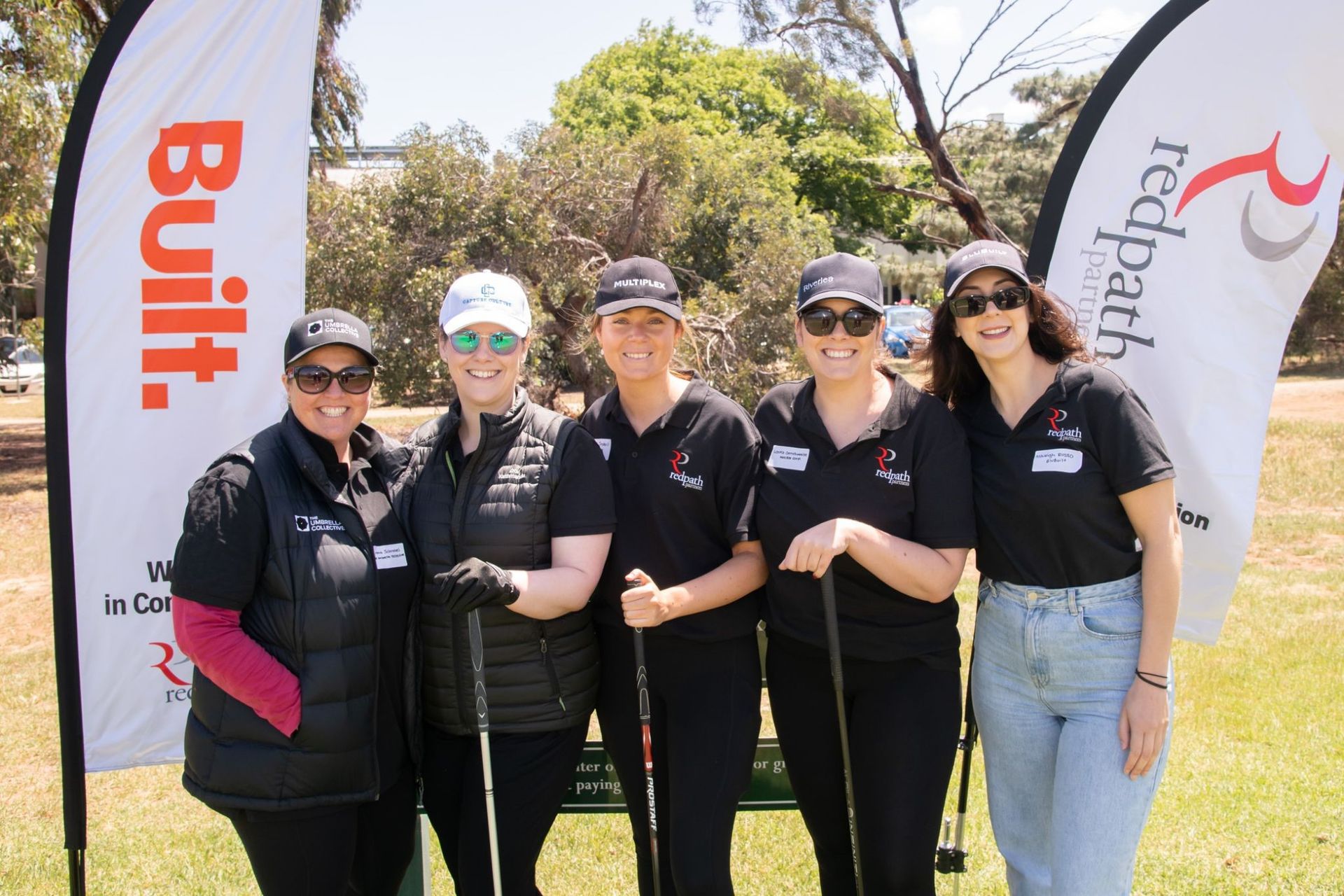 Five people wearing hats and matching black golf shirts pose outdoors between two branded 