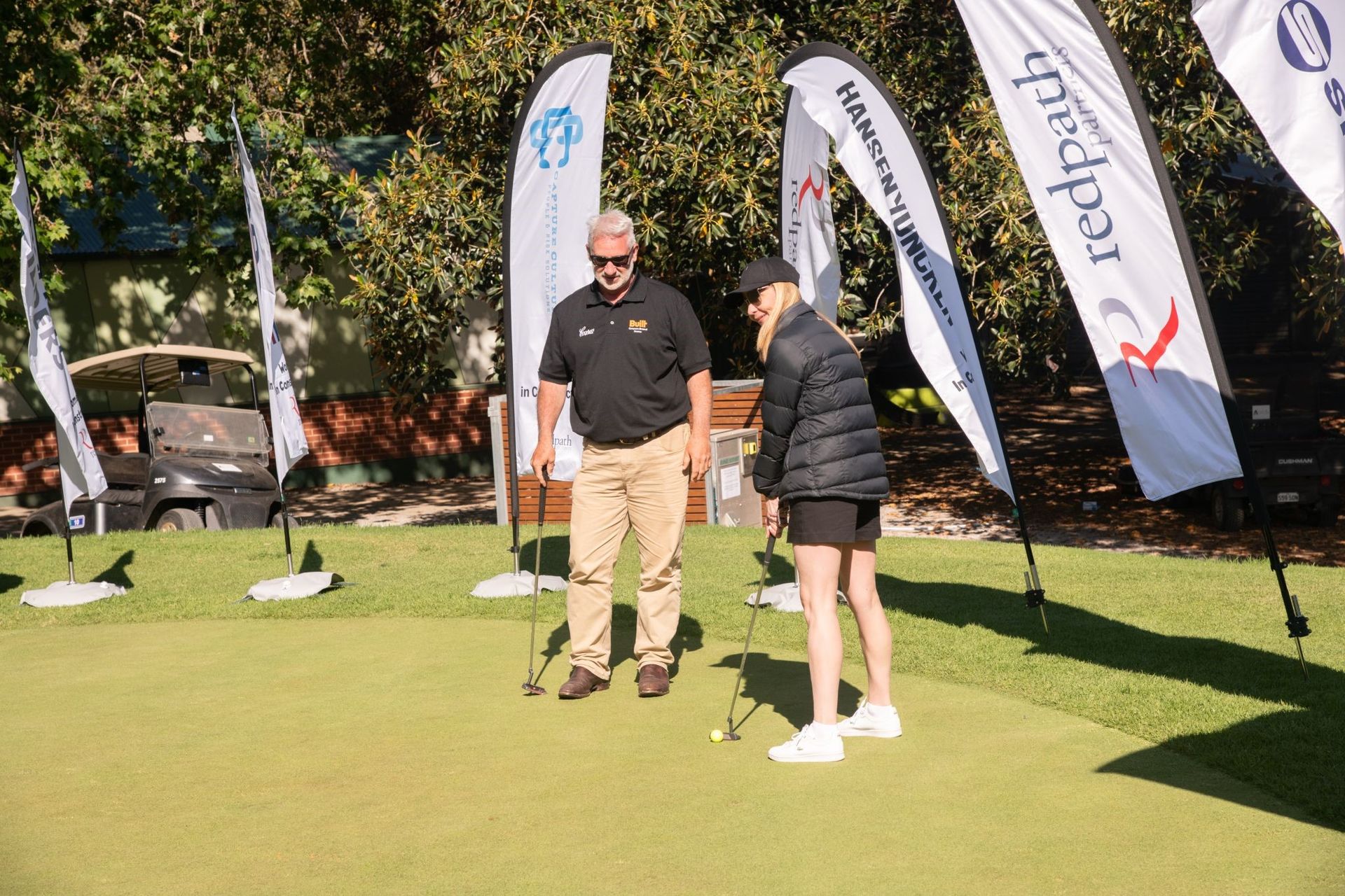 A person putting on a golf green while an instructor looks on, surrounded by promotional sponsor flags.