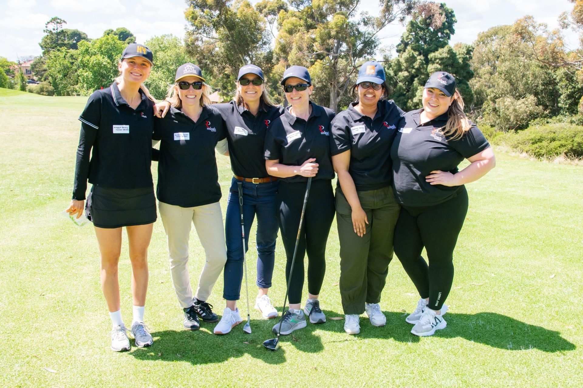A group of six people in matching black polo shirts and baseball caps posing together on a sunny golf course.