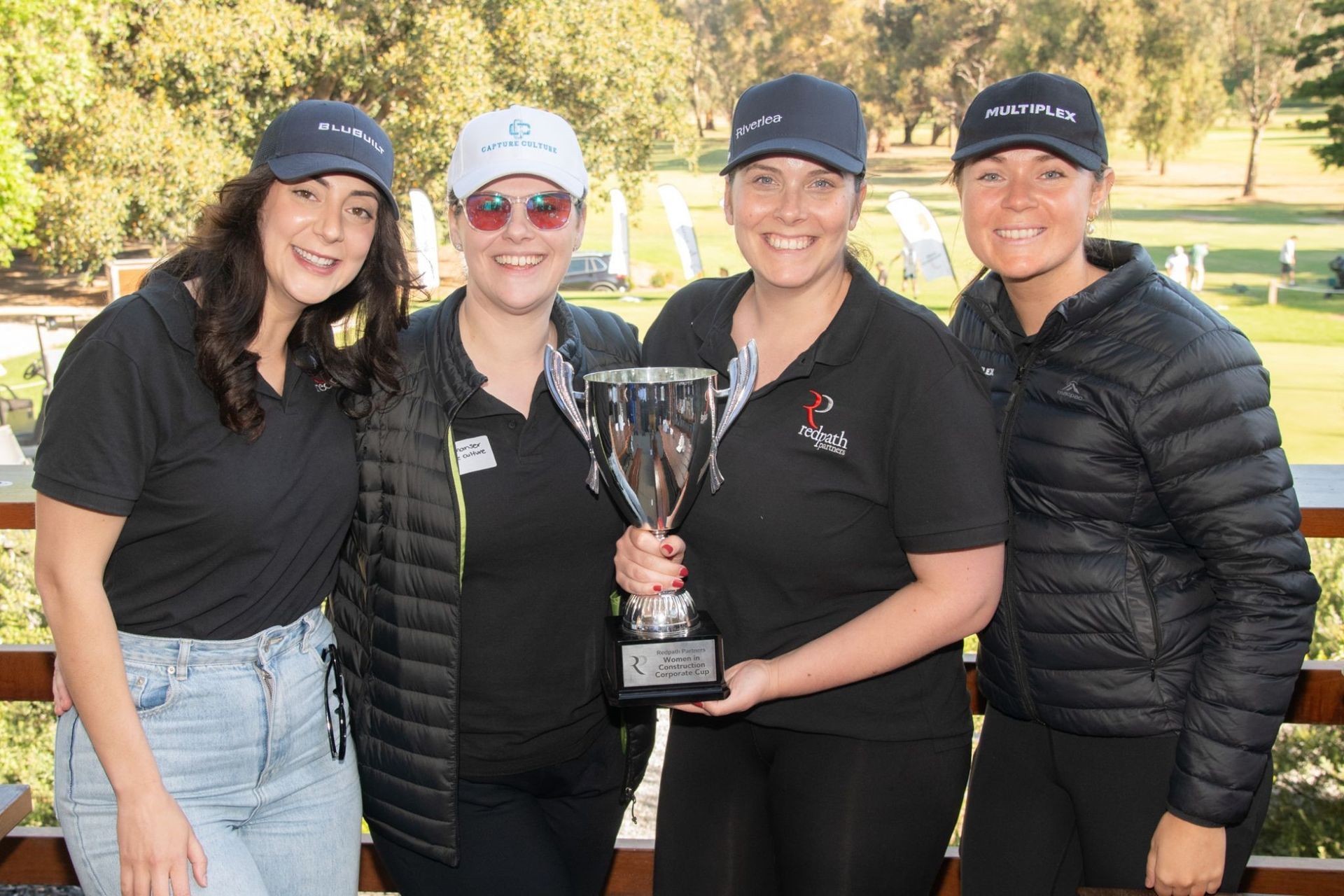 Four people smiling and holding a trophy outdoors in front of a golf course.
