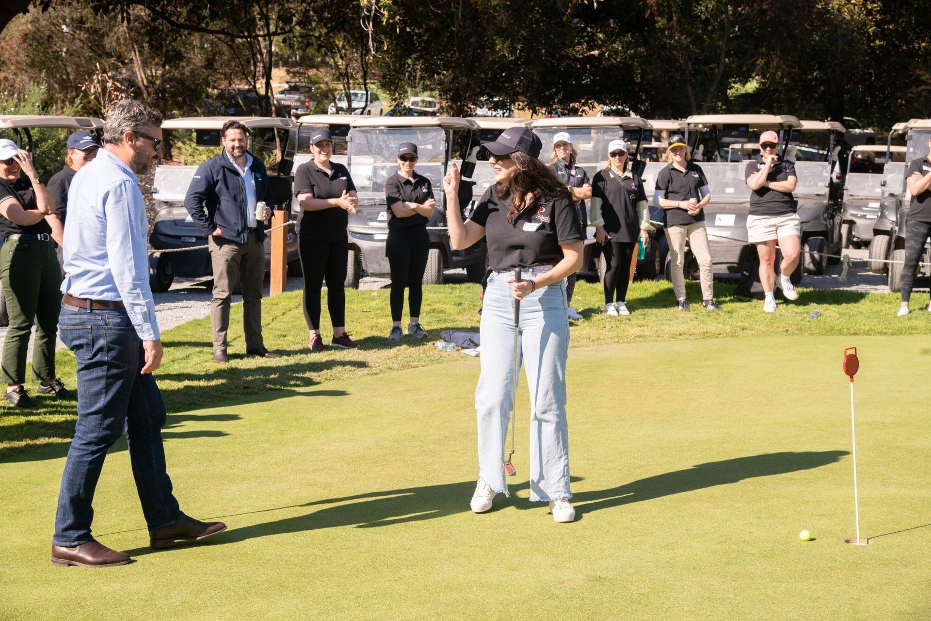 A person standing on a golf green with a putter, while a group of people watch from behind a row of parked golf carts.