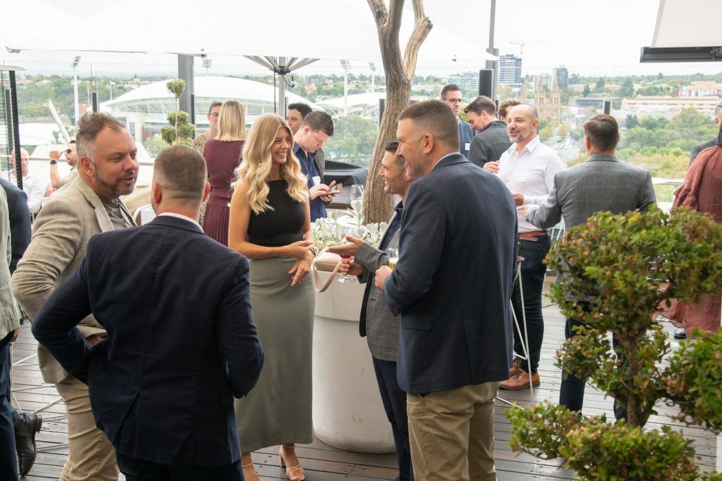 Group of people conversing at a professional outdoor networking event on a rooftop terrace with a scenic backdrop.
